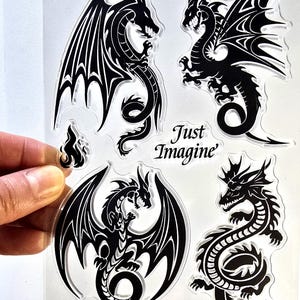 May include: A clear sheet featuring black dragon designs. The sheet includes four different dragon illustrations, each with detailed wings and serpentine bodies. The text "Just Imagine" is also present. The sheet is held by a hand.
