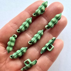 May include: A selection of small, green pea pod charms with white detailing. The charms are displayed on a hand, highlighting their miniature size and intricate design. These charms could be used for jewellery making or other craft projects.