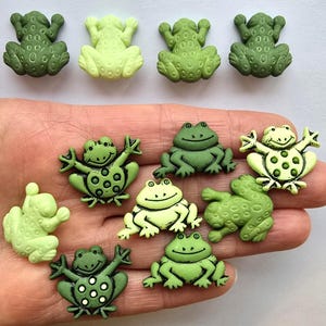 May include: A selection of green frog-shaped embellishments in various shades, including light and dark green. Some frogs have raised arms, while others are in a sitting position. The embellishments have a textured surface and are suitable for crafts.