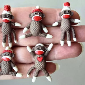 May include: Five miniature sock monkey figurines. Each brown monkey has white arms and legs, a red mouth, and a white hat with a red pom-pom. One monkey has a red heart on its chest. The monkeys are arranged on a hand.