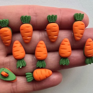 May include: A collection of orange carrot-shaped embellishments with green tops. The carrots are arranged on a light background. These small, decorative items are likely used for crafts or embellishments.