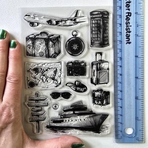 May include: Clear stamp set with travel-themed illustrations. Includes an aeroplane, suitcase, compass, camera, map, sunglasses, hat, backpack, signpost, and a cruise ship. A blue ruler is next to the stamps.