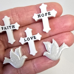 May include: A collection of white crosses with the words "FAITH", "HOPE", and "LOVE" in black lettering. Two pearlescent white dove figurines are also present. The items are likely religious-themed craft supplies or embellishments.