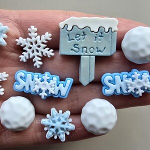 May include: A selection of winter-themed embellishments, including snowflakes, snowballs, and signs that read "Let it Snow". The snowflakes are white and light blue, and the snowballs are white. Ideal for craft projects.