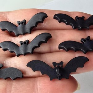 May include: Six small, matte black bat-shaped buttons. Each button has a detailed bat silhouette with outstretched wings and a small button shank on the back. Perfect for Halloween or craft projects.