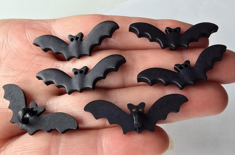 May include: Six small, matte black bat-shaped buttons. Each button has a detailed bat silhouette with outstretched wings and a small button shank on the back. Perfect for Halloween or craft projects.