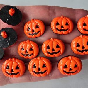 May include: A selection of Halloween-themed buttons and embellishments. The image shows several orange pumpkin-shaped buttons with black facial features, and black buttons with orange accents. These are likely used for crafting or sewing projects.