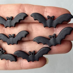 May include: Six black bat-shaped embellishments with outstretched wings. Each bat has two small eye holes and a small head. These decorative bats are ideal for Halloween or craft projects.
