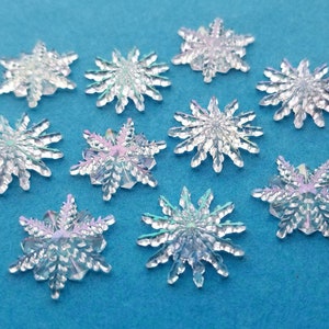 May include: Ten iridescent snowflake-shaped embellishments on a blue background. The snowflakes are made of a clear plastic material with a holographic finish.