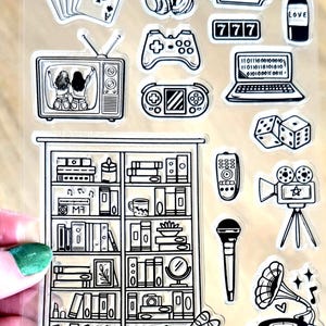 May include: Clear sticker sheet featuring black and white illustrations. The sheet includes playing cards, headphones, a ticket, a bottle with "LOVE", a TV, game controllers, a laptop, dice, a bookshelf, a remote, a microphone, a film camera, and a gramophone.