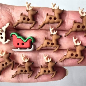 以下が含まれることがあります： クリスマスをテーマにしたボタンのコレクション。白い角を持つ茶色のトナカイと赤と緑のそりがあります。トナカイのボタンには茶色の斑点が2つあります。小さくて装飾的なボタンです。