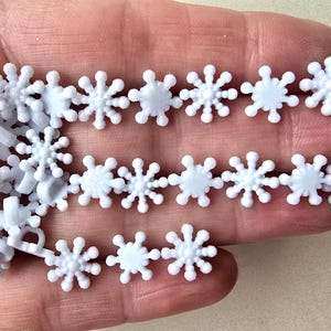 May include: A selection of small, white snowflake-shaped embellishments. The snowflakes are made of plastic and have a detailed, three-dimensional design. They are arranged in rows and scattered, ideal for crafts and winter-themed projects.