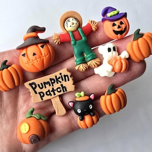 May include: A collection of small, colourful Halloween-themed polymer clay charms including pumpkins, a scarecrow, a ghost, a black cat in a pumpkin, and a tan sign with the text Pumpkin patch written in black, all featuring detailed textures.