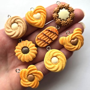 May include: A collection of eight miniature biscuit-shaped charms, each featuring a silver-toned metal jump ring. The charms display various textures and shapes, including round, heart, and flower designs, with central cream or brown coloured accents.