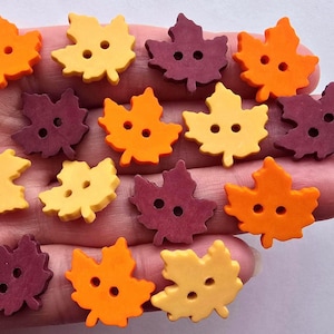 Peut inclure: Boutons en forme de feuilles d'érable dans des tons orange, jaune et bordeaux. Chaque bouton comporte deux trous pour la couture. Les boutons sont présentés sur un fond clair, pour des projets créatifs.