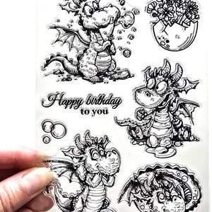 May include: Clear stamp set with cartoon dragon illustrations. Dragons are depicted in different poses, some with wings and scales. The set includes the text "Happy birthday to you".