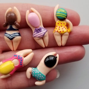 May include: Five miniature figurines of swimmers in various swimsuits. The figurines have tan bodies and arms raised above their heads. Swimsuits are in blue, purple, pink, yellow, and green with polka dots and stripes. The figurines are in a hand.