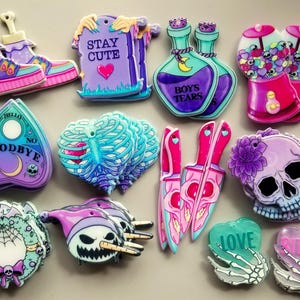 May include: A collection of 14 colourful resin charms with various designs, including a skull, a heart with ribs, a tombstone with the text "Stay Cute", a wreath, a gumball machine, a pair of boots, a pair of knives, a Ouija board, a bottle with the text "Boys Tears", and two hands with the text "Love" and "Rites".
