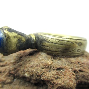 Può includere: Anello antico in ottone con una gemma blu vibrante. L'anello presenta dettagli incisi e una finitura ossidata, conferendogli un aspetto vintage. La gemma è di un blu intenso, aggiungendo un tocco di colore. Probabilmente un pezzo d'epoca.