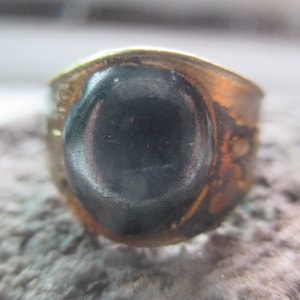 May include: A vintage ring with a dark, possibly black, oval-shaped stone set in a tarnished gold-toned metal band. The ring's band shows signs of wear, suggesting it is an antique or vintage piece. The stone is smooth and polished.