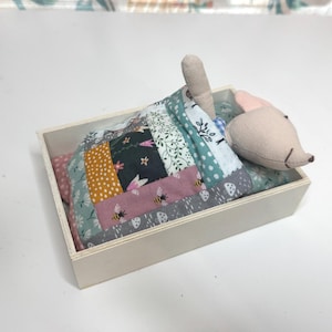 May include: A small, wooden box contains a miniature bed with a patchwork quilt and a stuffed mouse. The quilt features various fabric patterns, including floral, polka dots, and bee motifs. The mouse has a light pink nose and ears.