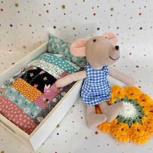 Puede incluir: Un juguete de peluche con forma de ratón con orejas rosas y vestido de cuadros vichy azul y blanco, sentado junto a una pequeña cama de madera. La cama tiene una colcha de retazos con motivos de flores, abejas y setas, y una almohada a juego.