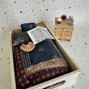 May include: A miniature bed set in a wooden tray featuring a patchwork quilt with navy, brown, and houndstooth patterns. Accessories include a tiny newspaper, a berry tart on a woven plate, a toy ship, a nightstand with a clock, and a coffee cup.
