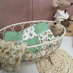May include: A miniature white metal crib holds a patchwork quilt with green polka dots and floral patterns. A small, beige crocheted blanket and a cream-colored doily are nearby. Two stuffed mice are in the crib and on a wooden rocking horse.