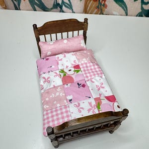 May include: A miniature wooden bed with a pink patchwork quilt and pillow. The quilt features various pink floral and gingham patterns. The bed frame is a dark brown color.