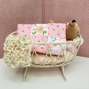 May include: A miniature crib with a beige metal frame holds a patchwork quilt with pink stripes and floral patterns. A small, brown teddy bear rests on the pillow. A cream-colored knitted blanket is draped over the side.