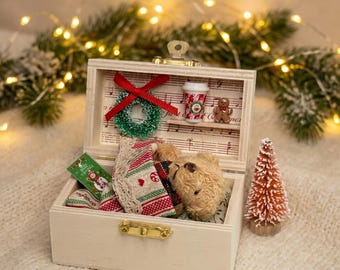 May include: A light-colored wooden box containing a miniature teddy bear tucked under a patterned fabric blanket. The interior lid features a small green wreath with a red bow, a miniature coffee cup, and a tiny gingerbread man figurine.