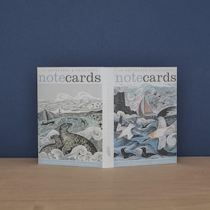 May include: Two boxes of notecards and envelopes are shown. The notecards have illustrations of a seal and a sailboat on the sea. The text "10 Notecards & Envelopes" and "notecards" are printed on the front. The background is dark blue.