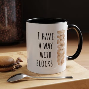 May include: White ceramic mug with a black interior and handle. The mug features the text "I HAVE A WAY WITH BLOCKS." and a graphic of stacked wooden blocks. A spoon and a pastry are on a wooden surface.