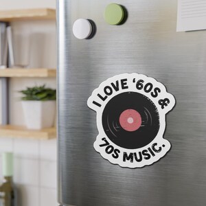 May include: A white magnet with a black vinyl record design and the text "I LOVE '60s & 70s MUSIC." The record has a pink center. The magnet is on a silver refrigerator door with other magnets.