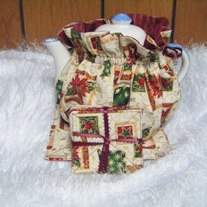 May include: A white teapot with a Christmas-themed fabric cover. The cover is decorated with red poinsettias, green holly, and gold stars on a cream background. The teapot is sitting on a white fluffy surface.