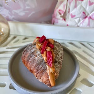 Könnte beinhalten: Ein Croissant auf einem grauen Teller, gefüllt mit einer hellbraunen Creme und belegt mit roten rechteckigen Stücken. Das Gebäck liegt auf einem weißen Tablett, mit einer rosa Blumenvase und einer rosa herzförmigen Tasche im Hintergrund.