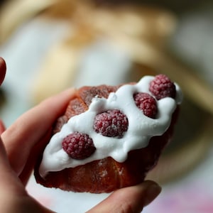 Könnte beinhalten: Nahaufnahme eines Gebäckstücks, möglicherweise eines Croissants, das mit weißem Zuckerguss und frischen Himbeeren belegt ist. Das Gebäck wird in einer Hand gehalten, mit unscharfem Hintergrund. Die Himbeeren sind leuchtend rot und kontrastieren mit dem weißen Zuckerguss.
