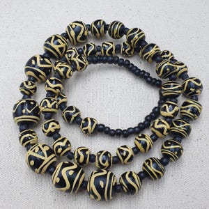 Op de afbeelding: Een lange ketting gemaakt van zwarte en gele houten kralen. De kralen zijn rond en hebben een geometrisch patroon.