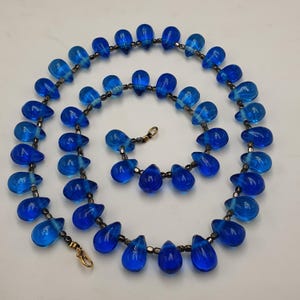May include: A necklace featuring teardrop-shaped blue glass beads. The beads are strung together with small, dark gray beads and a gold-colored clasp. The necklace is displayed on a white surface.