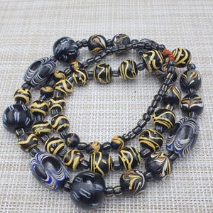 Könnte beinhalten: Eine Halskette aus schwarzen und gelben Glasperlen mit verschiedenen Mustern. Die Perlen sind rund und haben eine glänzende Oberfläche.