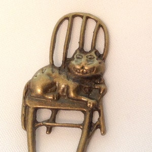 May include: A gold-toned metal charm of a cat sitting in a chair. The cat has a mischievous grin and is sitting with its paws crossed.