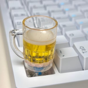 May include: A small beer mug, filled with golden liquid and white foam, sits on a white mechanical keyboard. The clear mug has a handle and is positioned between the keys, adding a playful touch to the setup.