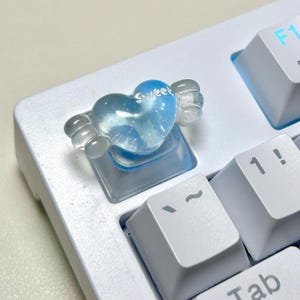 Pode incluir: Uma tecla de teclado personalizada em forma de coração com asas azuis translúcidas e transparentes. A palavra "Sweet" está escrita no coração. A tecla está em um teclado branco.