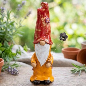Può includere: Statuetta di gnomo da giardino in ceramica con un cappello rosso alto e un cappotto arancione. Lo gnomo ha una lunga barba bianca, un naso piccolo e tiene in mano un fiore nero. Un piccolo fiore viola decora il cappello. La statuetta è su una superficie di legno.