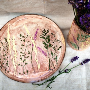 Könnte beinhalten: Ein handgefertigtes Keramik-Set aus Teller und Vase mit floralem Design. Der Teller hat einen Durchmesser von ca. 25 cm und zeigt grün, gelb und violett bemalte Pflanzen. Die Vase enthält lila Lavendel.