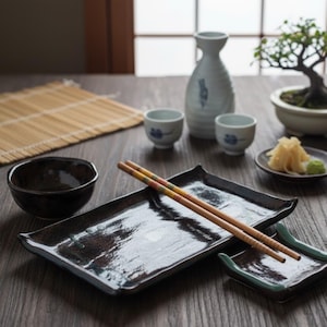 Può includere: Un'ambientazione giapponese per sushi con una ciotola marrone scuro, due piatti rettangolari e un paio di bacchette. Sono presenti anche tazze da sake, una bottiglia di sake e un piccolo bonsai. I piatti sono lunghi circa 20 cm.