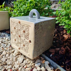 Può includere: Una fioriera quadrata in ceramica beige con manico grigio. La fioriera presenta un motivo floreale su un lato e piccoli fori. La fioriera è appoggiata su un letto di piccole rocce, con piante verdi sullo sfondo.