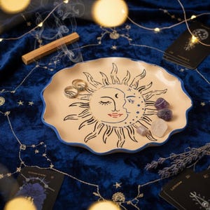 Könnte beinhalten: Eine beige Keramikschale mit einem Sonnen- und Monddesign, schwarzen Umrissen und einem blauen Rand. Zwei goldene Ringe und Kristalle befinden sich auf der Schale, mit Tarotkarten und einem Räucherstäbchen aus Holz in der Nähe. Die Schale steht auf einer blauen Samtoberfläche.
