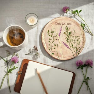 Op de afbeelding: Een serene stilleven met een kopje thee, een brandende kaars en een decoratieve plaat met bloemmotieven en de tekst "Let it be...". Een notitieboek, een potlood en bloemen maken de scène compleet.