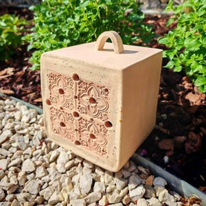 Könnte beinhalten: Ein quadratisches Insektenhaus in Terrakotta-Farbe mit einem Griff oben. Die Vorderseite weist ein geprägtes Blumendesign mit kleinen Löchern auf. Das Haus steht auf einem Bett aus kleinen, weißen Steinen, mit grünem Laub im Hintergrund.